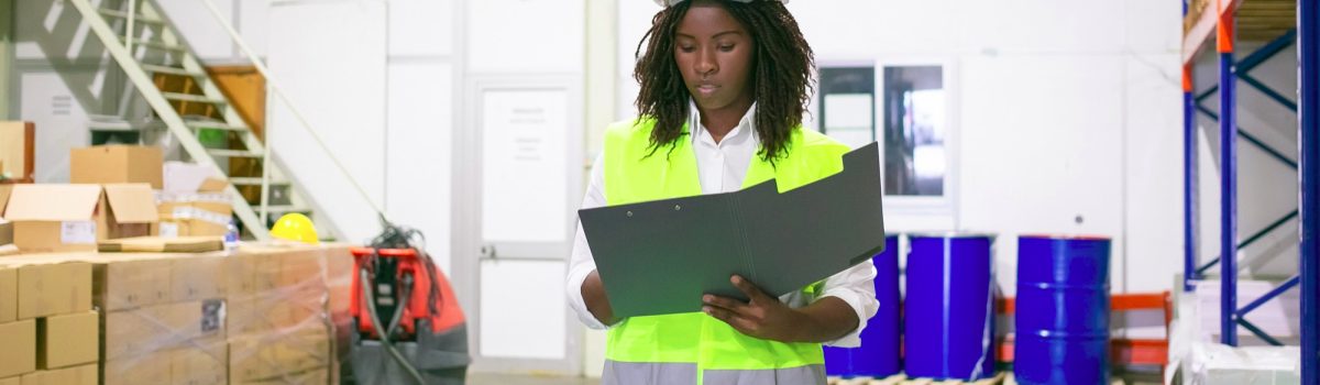 focused-female-logistic-employee-hardhat-safety-vest-walking-warehouse-carrying-open-folder-looking-through-document-copy-space-front-view-labor-inspection-concept